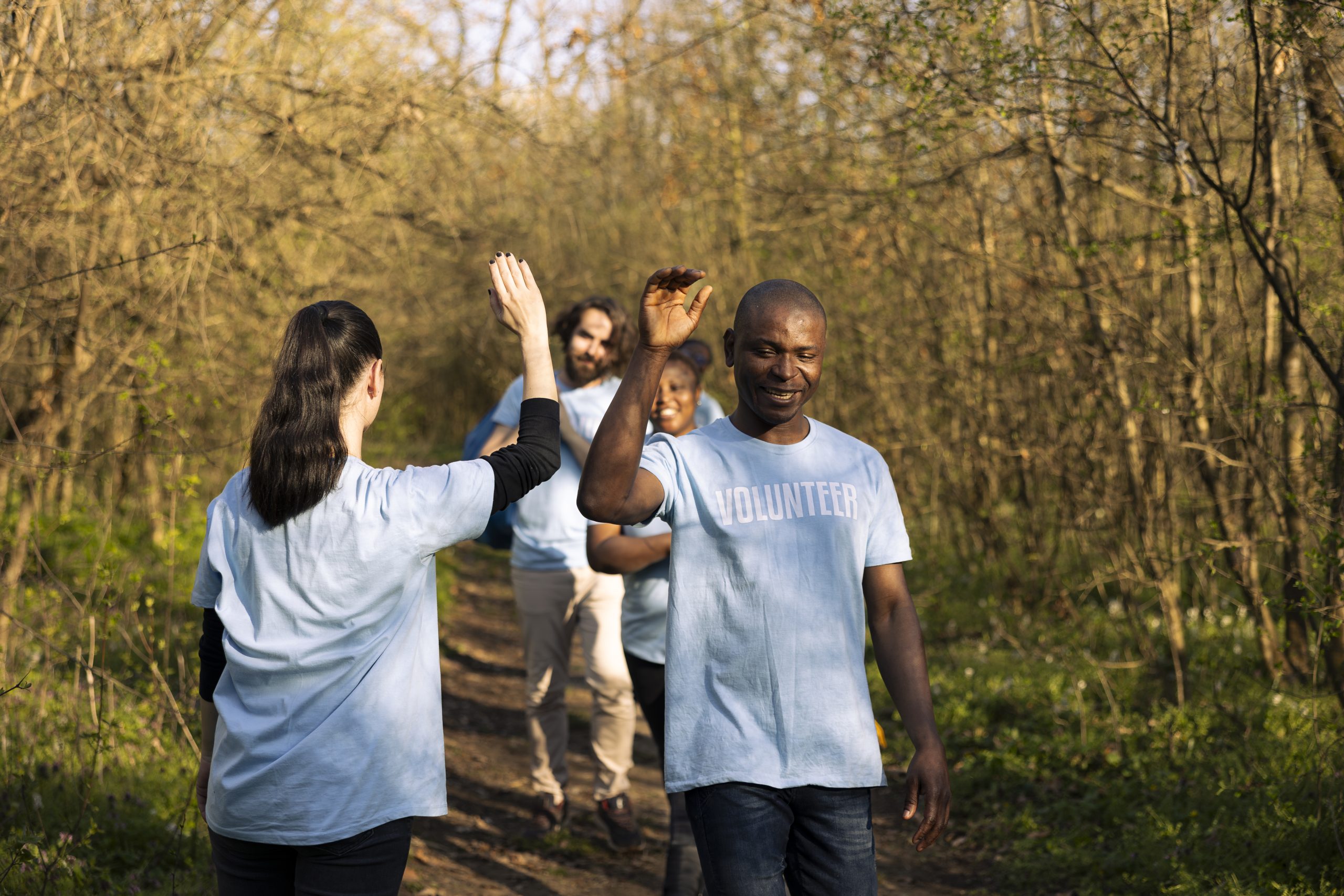 Cheerful satisfied activists sharing high five with each other in the woods, finishing to tidy up the forest ecosystem against pollution. Pleased volunteers showing praise after a job well done.