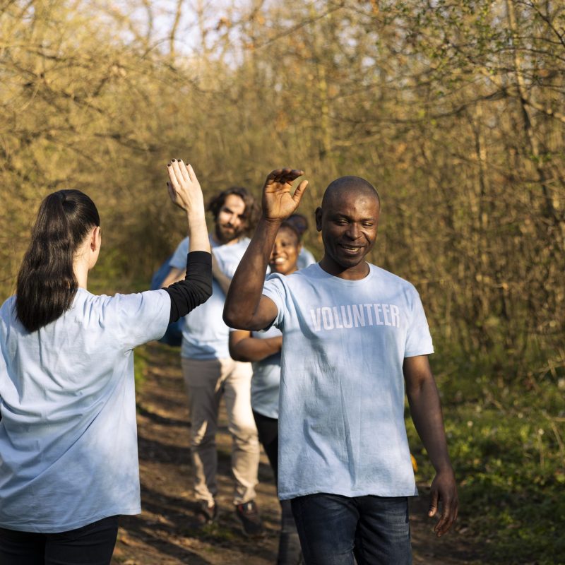 Cheerful satisfied activists sharing high five with each other in the woods, finishing to tidy up the forest ecosystem against pollution. Pleased volunteers showing praise after a job well done.