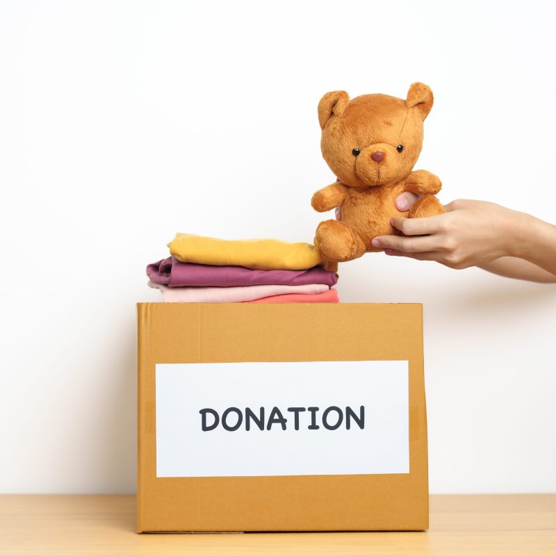 Kid Donation, Charity, Volunteer, Giving and Delivery Concept. Hand holding Bear doll and Clothes into Donation box at home for support and help poor, refugee and homeless people. Copy space for text